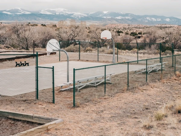 Outdoor basketball court with chain-link fence in desert setting, distant snow-capped mountains under cloudy sky, benches, and bare trees.