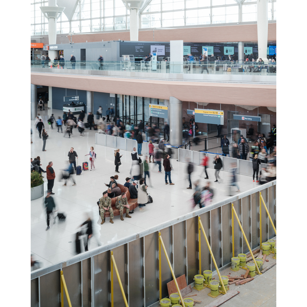 Busy airport terminal interior with blurred travelers, uniformed officers seated on a bench, and temporary flood barriers in the foreground.