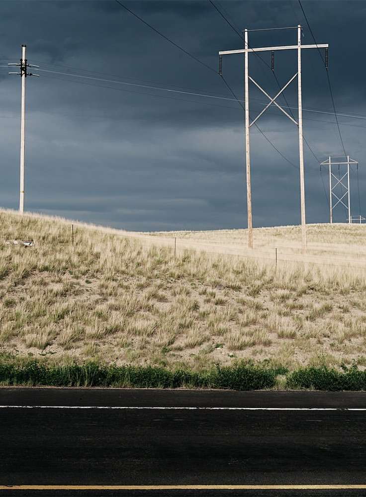 Landscape view of power lines on a grassy hill against a dark, overcast sky, with a dark asphalt road in the foreground.