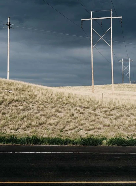 Landscape view of power lines on a grassy hill against a dark, overcast sky, with a dark asphalt road in the foreground.