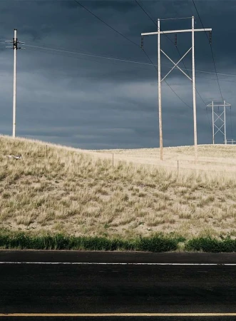 Landscape view of power lines on a grassy hill against a dark, overcast sky, with a dark asphalt road in the foreground.