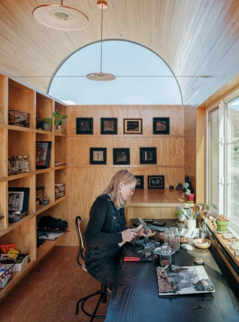 Woman crafting at desk in minimalist studio. Interior with built-in shelves, wooden walls, arched window, and pendant lights, bathed in natural light.