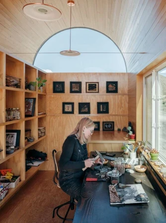 Woman crafting at desk in minimalist studio. Interior with built-in shelves, wooden walls, arched window, and pendant lights, bathed in natural light.