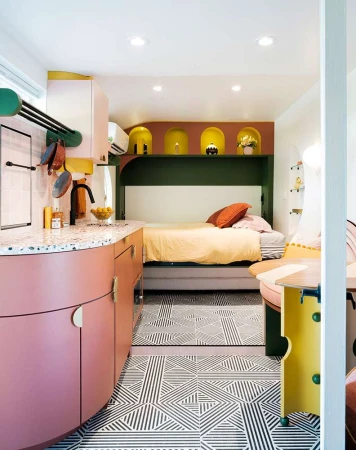 Interior shot of a compact kitchen and living space featuring pink cabinets, patterned floors, and a neatly made bed.