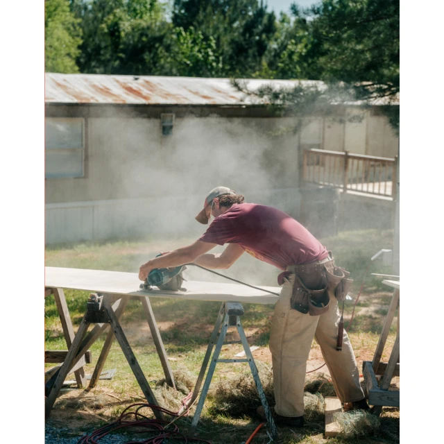 Man wearing a cap and toolbelt cuts wood with a power saw on a wooden sawhorse outside, creating sawdust with a house in the background.