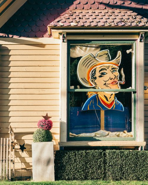 Whimsical exterior view of a yellow house with a kitschy cowboy illustration in the window, lush green hedge, and decorative flower pot.