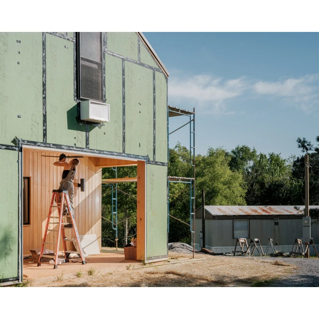 Exterior construction site features a man painting interior wood framing, standing on a ladder near a house under construction on a sunny day.