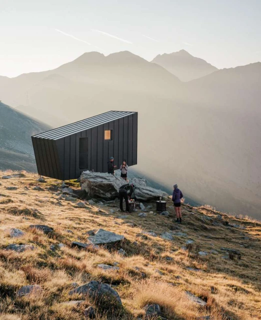 Modern mountain hut nestled in a scenic range, surrounded by grassy slopes, rocks, and a group of people enjoying the landscape and architecture.