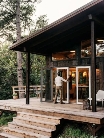 Adult male opening sliding glass doors to a dark wood cabin with a wood deck, stairs, outdoor seating, and forest background.