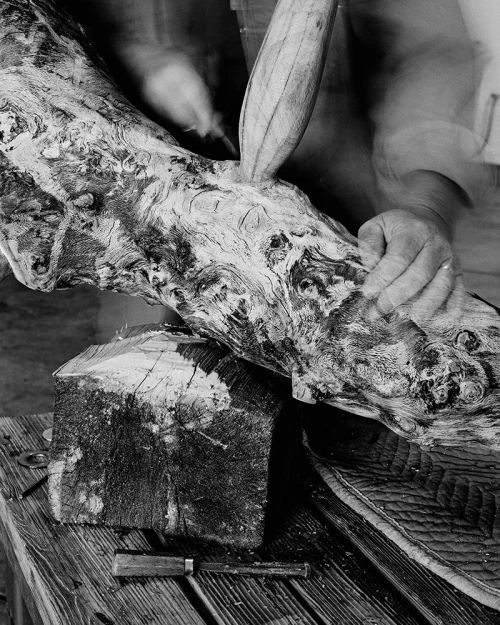 Black and white image of a woodworker carving a piece of wood with a chisel, set on a wooden table with additional tools.