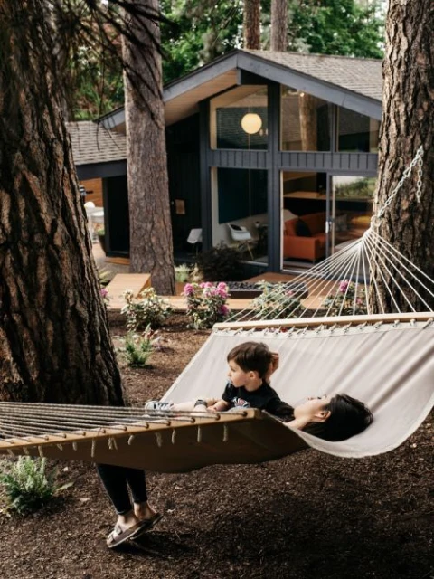 A child and an adult are resting in a hammock outside a modern house surrounded by trees, creating a peaceful and family-friendly scene.