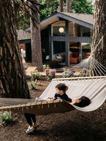 A child and an adult are resting in a hammock outside a modern house surrounded by trees, creating a peaceful and family-friendly scene.