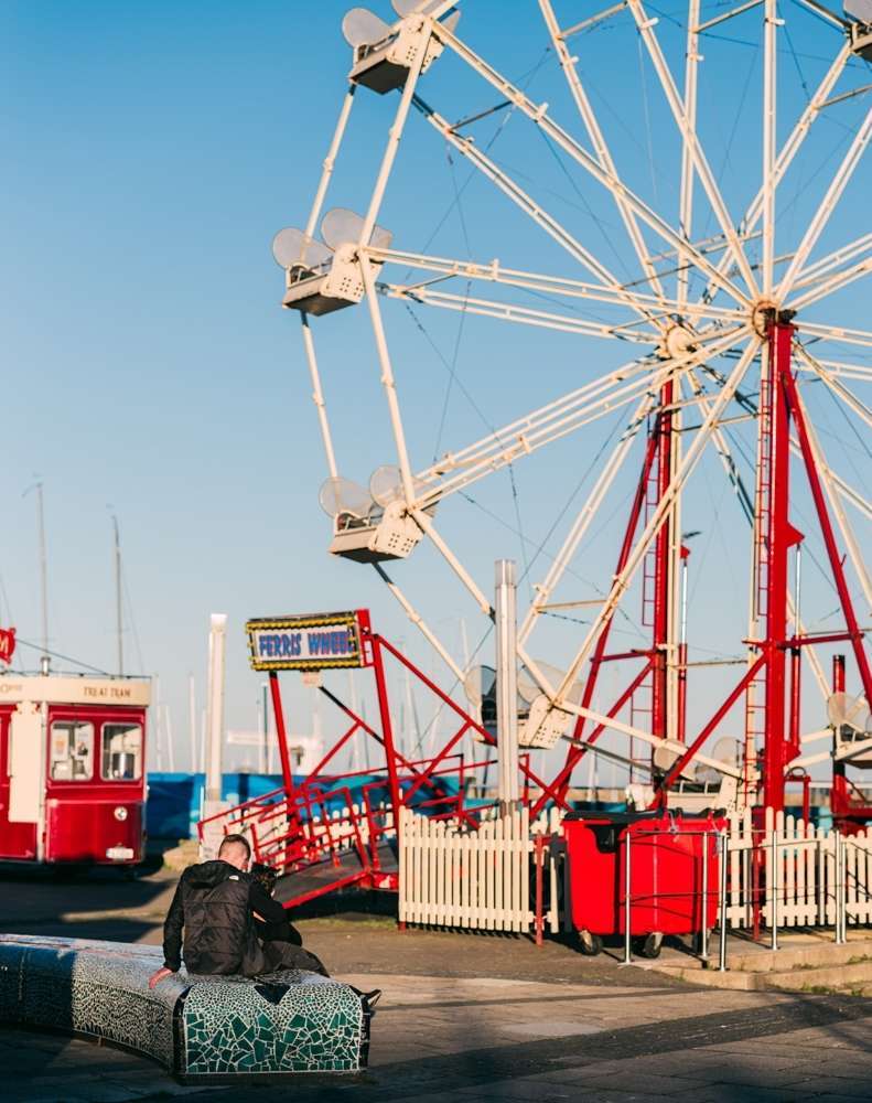 Adult male seated on a mosaic bench near a Ferris wheel with red and white accents on a clear day with other structures in the background.
