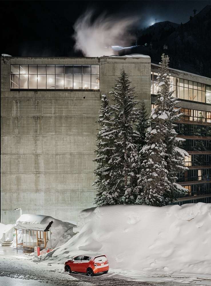Night scene featuring a modern concrete building with illuminated windows, snow-covered trees, a red car, and a snow-covered landscape.