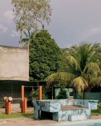 Outdoor courtyard with a swimming pool, painted concrete structures, palm trees, and an overcast sky by Benjamin Rasmussen