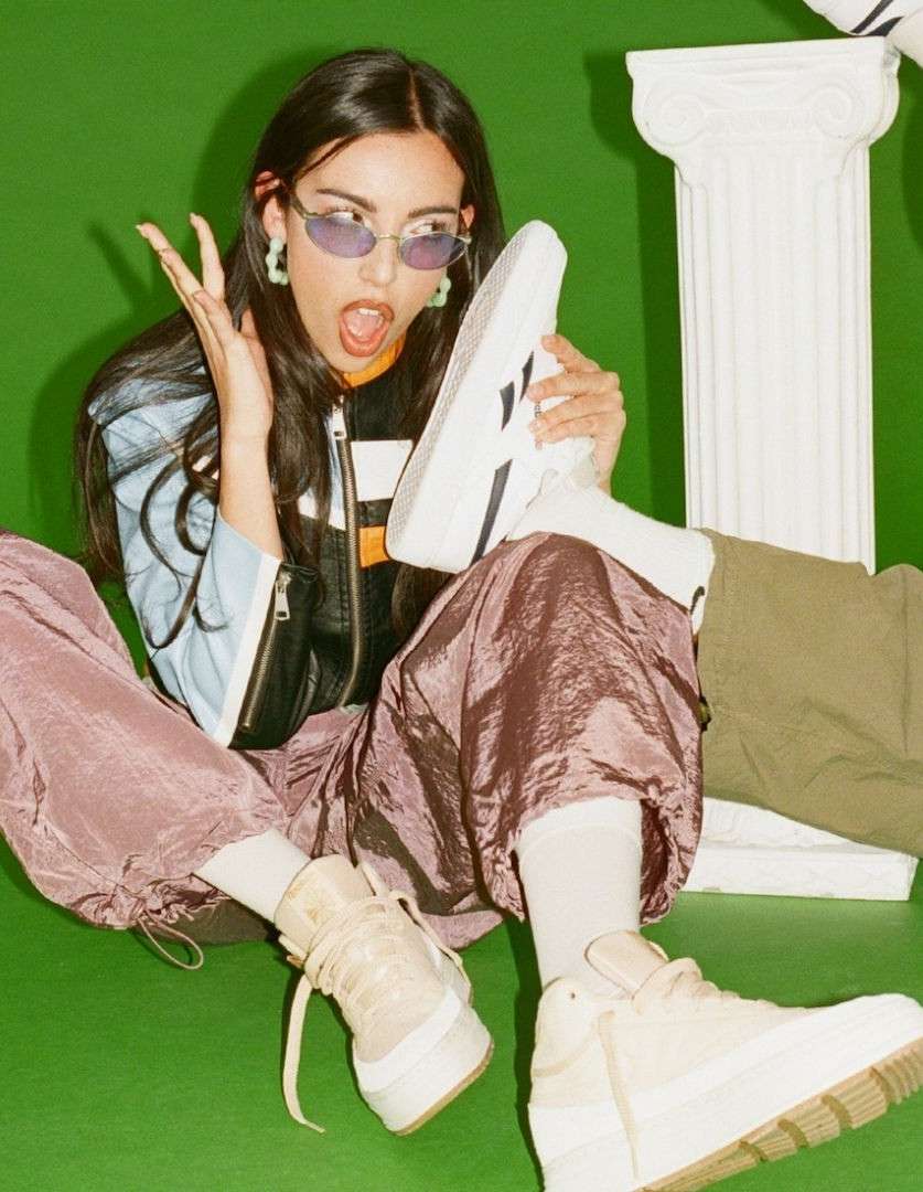Woman seated on a green background wearing purple pants and oval glasses, holding a white sneaker with a white column in the background.