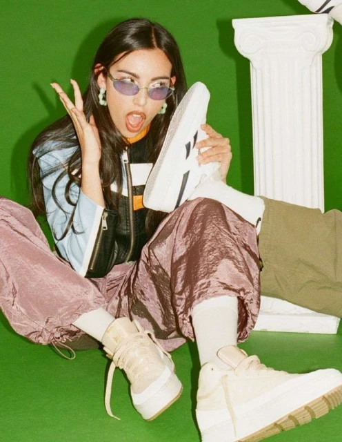 Woman seated on a green background wearing purple pants and oval glasses, holding a white sneaker with a white column in the background.
