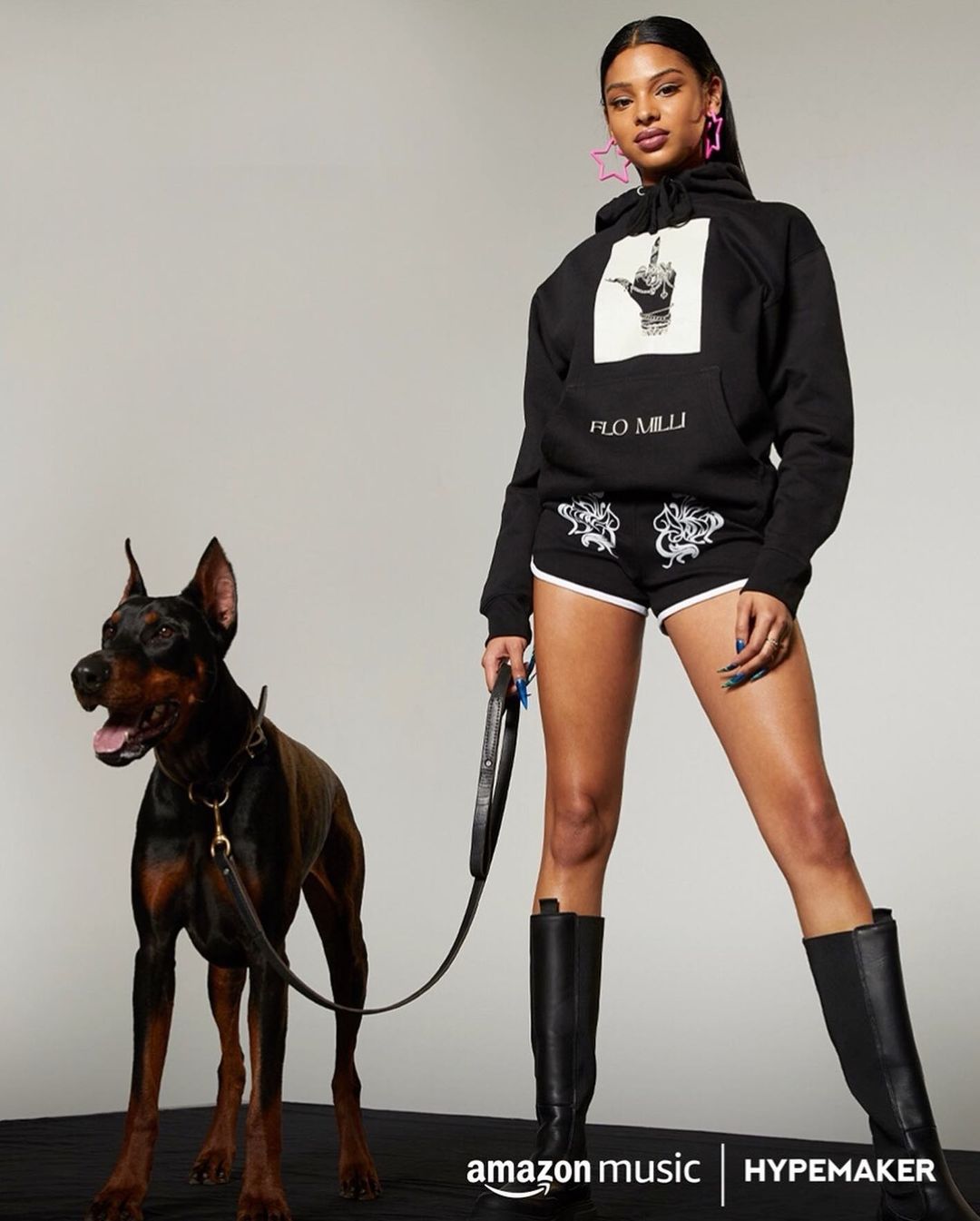 Adult female poses next to a black doberman pinscher wearing a collar. The model wears a black hoodie, shorts, and high leather boots.