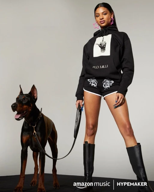 Adult female poses next to a black doberman pinscher wearing a collar. The model wears a black hoodie, shorts, and high leather boots.