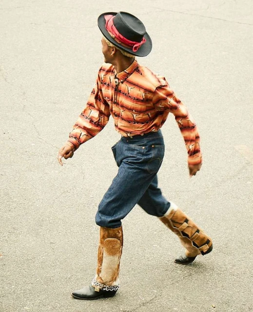 Man in stylish hat, patterned shirt, jeans, and ornate boots walks in a profile outdoor shot on a light-gray surface.