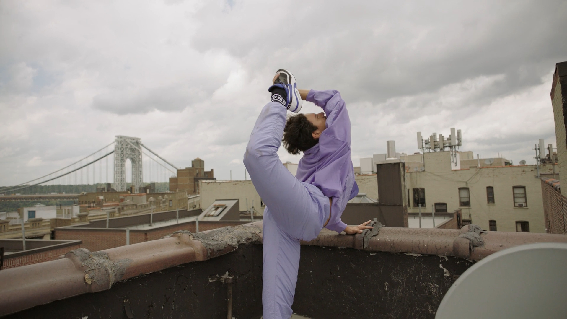 Female yogi strikes a standing bow pose on an urban rooftop with a city skyline and suspension bridge in the background on a cloudy day.