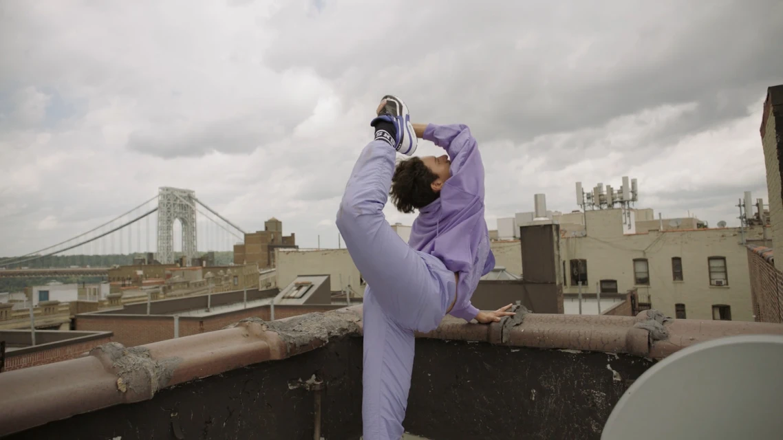 Female yogi strikes a standing bow pose on an urban rooftop with a city skyline and suspension bridge in the background on a cloudy day.