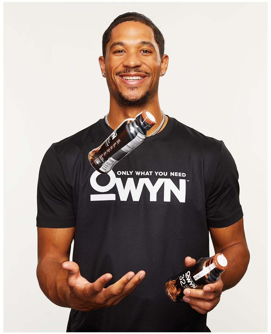 Smiling man in a black t-shirt juggling OWYN protein drinks in a bright indoor setting, promoting fitness and healthy living.