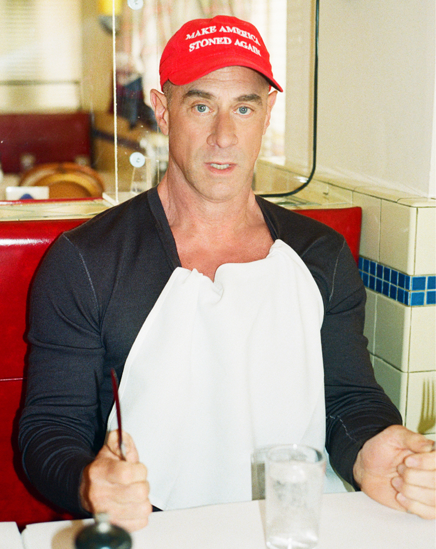 A man in a red cap sitting at a diner table with a bib on, holding silverware, and looking directly at the camera.