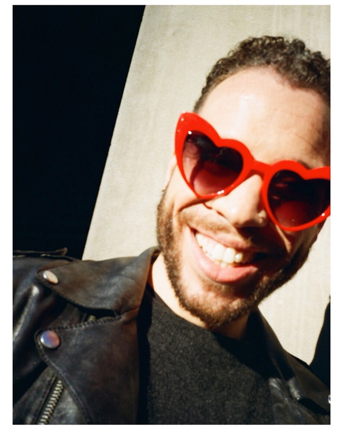 Smiling adult man wearing heart-shaped sunglasses and a leather jacket in front of a concrete wall with a black area to the left.