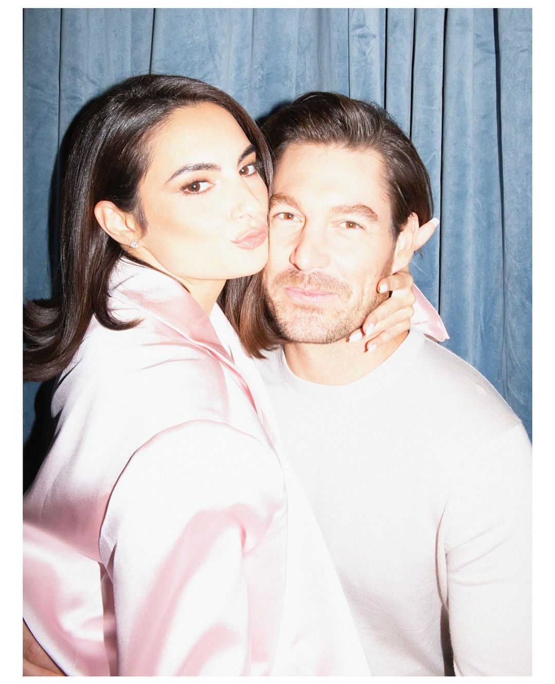 A man and a woman pose closely together against a blue curtain. The woman wears pink satin and the man a light gray shirt.