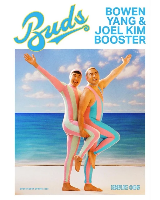 Two men in matching striped bodysuits pose on a beach against a blue sea and sky for a magazine cover featuring Bowen Yang and Joel Kim Booster.