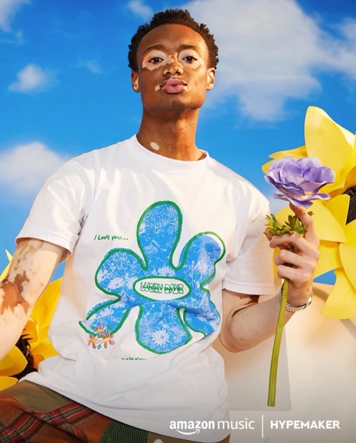 Young man with vitiligo poses with a flower in a light-filled portrait featuring a printed t-shirt, a blue sky, and large yellow flowers.