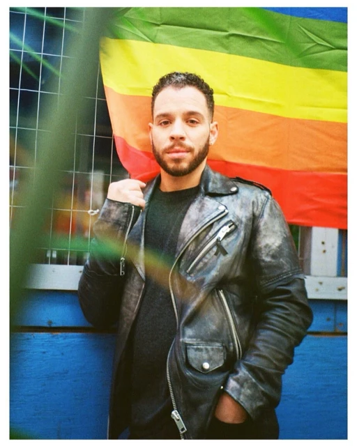 Adult male with a beard poses outdoors wearing a black leather jacket, with a rainbow flag draped over his shoulder, standing against a blue wall.