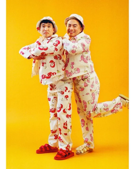 Two Asian adults in matching cherry-patterned outfits and bucket hats pose indoors against a bright yellow backdrop with a fun vibe.
