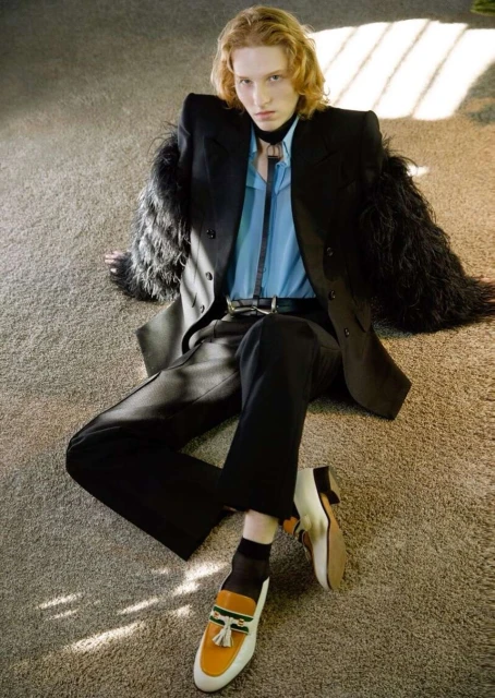 Fashion shot of a young male model with shoulder-length wavy hair in a black blazer and black dress pants sitting on a carpeted floor.