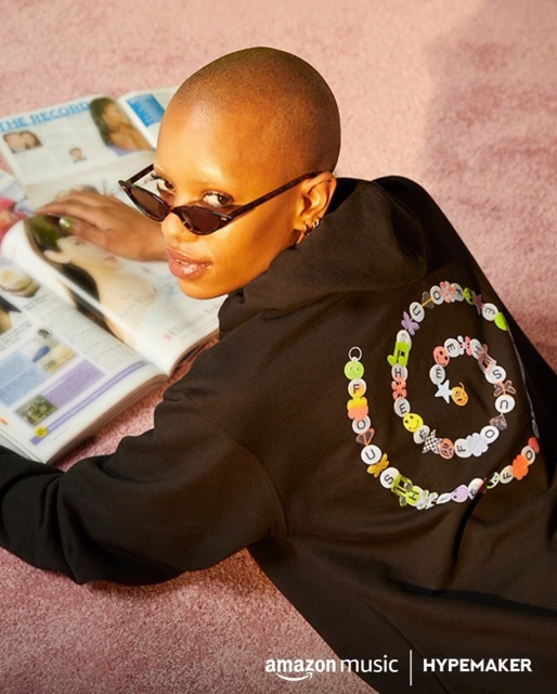 Young adult with sunglasses and short hair poses with black hoodie, magazine, and Amazon Music HYPEMAKER logo on pink carpet.