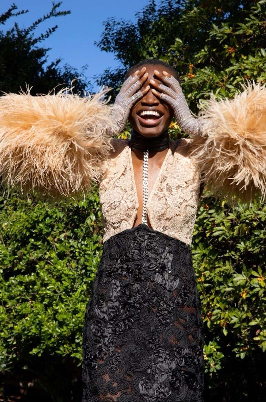 A cheerful black woman in nature covers her eyes with her hands, wearing a lacy dress with dramatic feathered sleeves in warm sunlight.