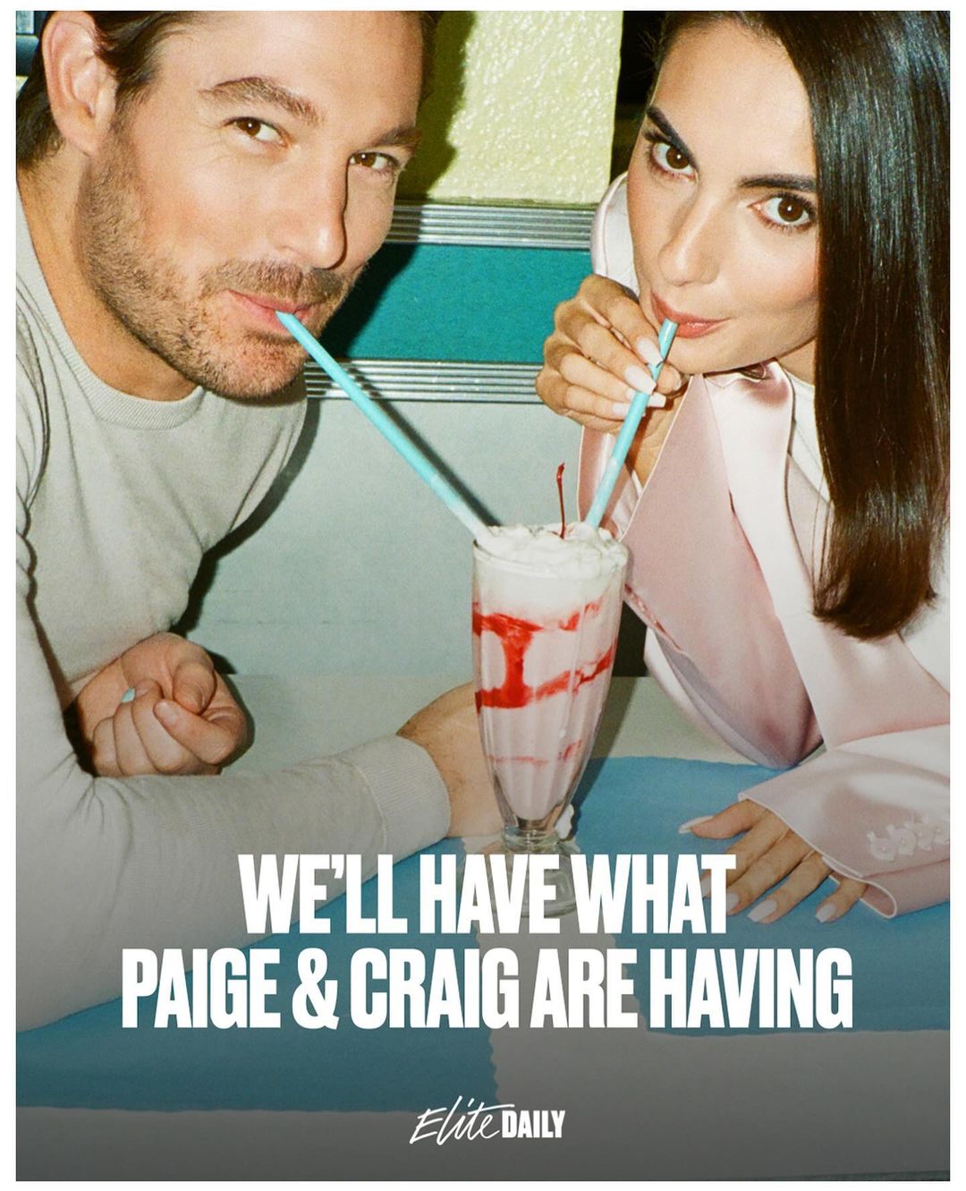 Attractive couple shares a pink milkshake with a cherry on top in an indoor setting, both sipping from blue straws, well-lit scene.