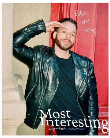 Man in a black leather jacket poses with hand raised to his forehead in a half salute, standing next to a red door and a stone pillar.