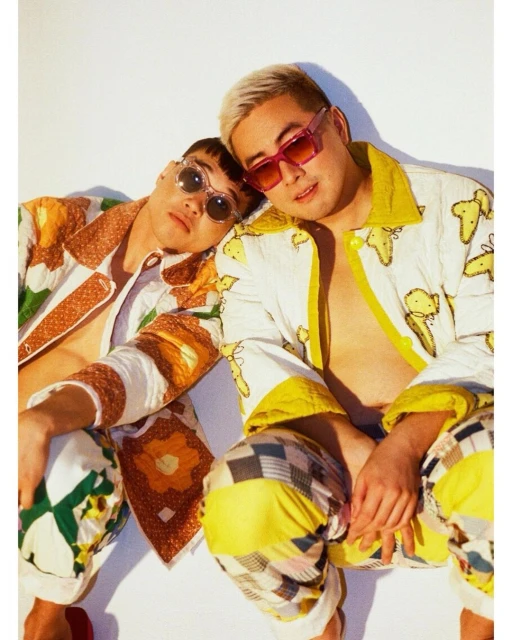 Two stylish young men pose in patterned jackets and pants against a white background, wearing sunglasses in an indoor portrait.
