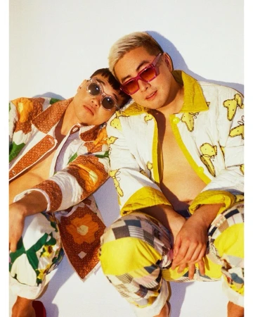 Two stylish young men pose in patterned jackets and pants against a white background, wearing sunglasses in an indoor portrait.