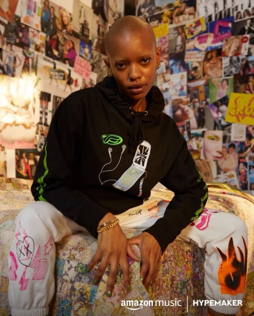 A young adult woman with a shaved head is sitting on a bed wearing a black hoodie and white sweatpants in an indoor setting.