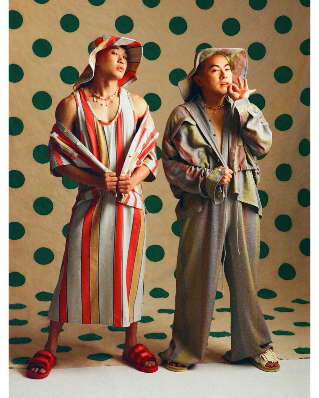 Two fashion models in striped outfits and hats pose against a polka dot background in a studio setting, showcasing vibrant and unique style.