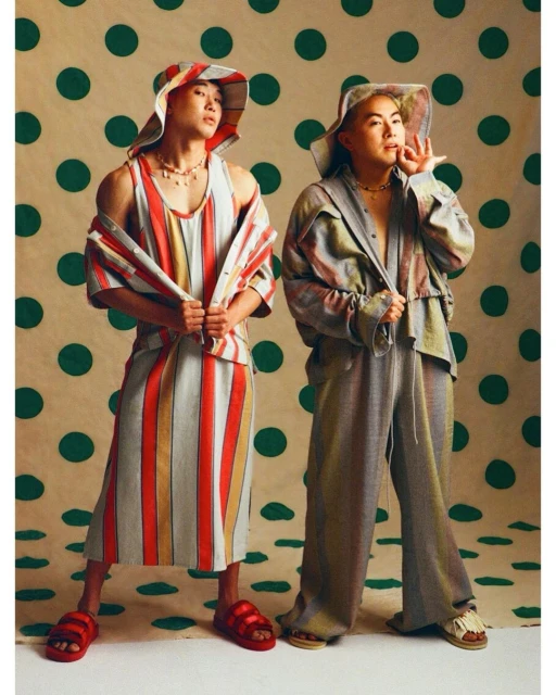 Two fashion models in striped outfits and hats pose against a polka dot background in a studio setting, showcasing vibrant and unique style.