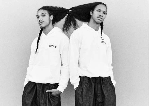 Two young adult men standing back-to-back in matching white hoodies and dark pants, posing in a studio with long, dark, styled hair.