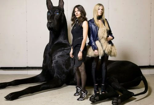 Two adult women pose next to a large, black Great Dane statue in an indoor setting with simple backgrounds and modern fashion.