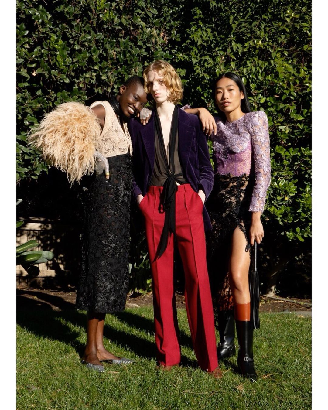 Three diverse fashion models pose together in an outdoor setting with lush green foliage, showcasing stylish eveningwear and textures.