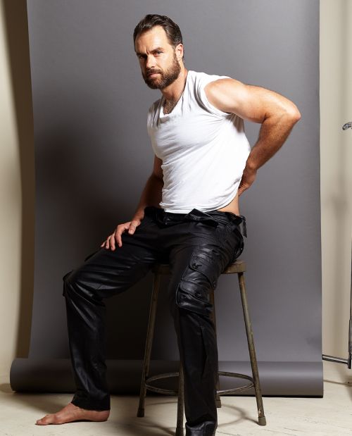 An adult male with a beard poses in a photo studio, wearing a white t-shirt and black leather pants while sitting on a metal stool.
