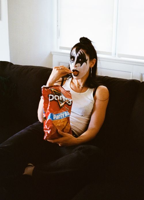 Woman with KISS-style makeup sits on a couch while eating Doritos; white walls, black hair, white tank top, and a red Doritos bag visible.