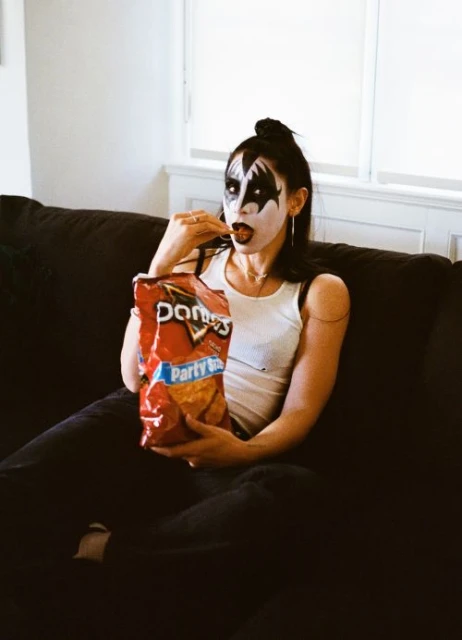 Woman with KISS-style makeup sits on a couch while eating Doritos; white walls, black hair, white tank top, and a red Doritos bag visible.